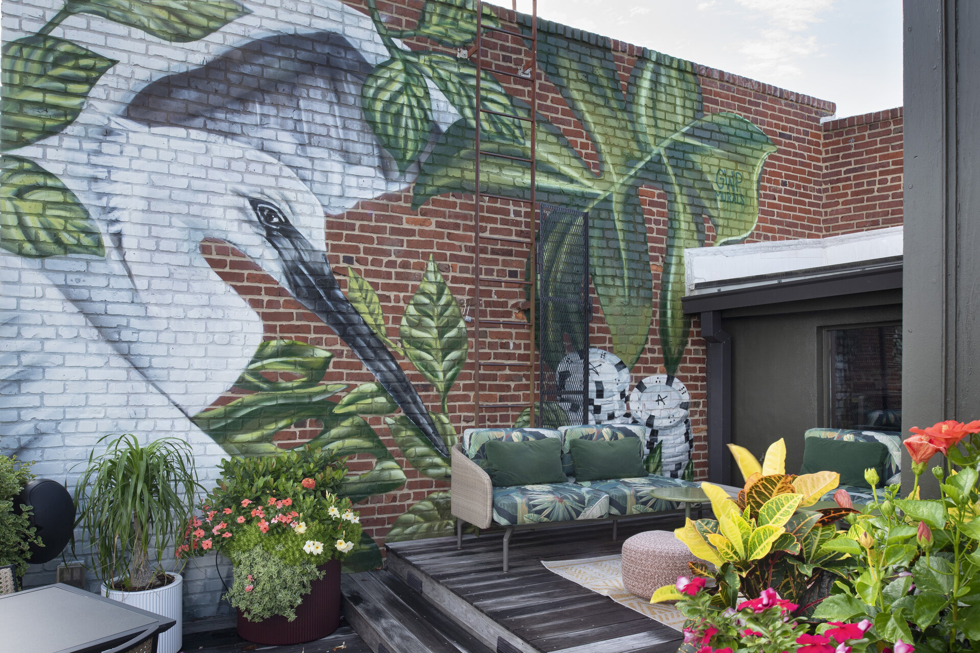 A mural of a bird and plants on a brick wall in an outdoor seating area with plants and flowers.