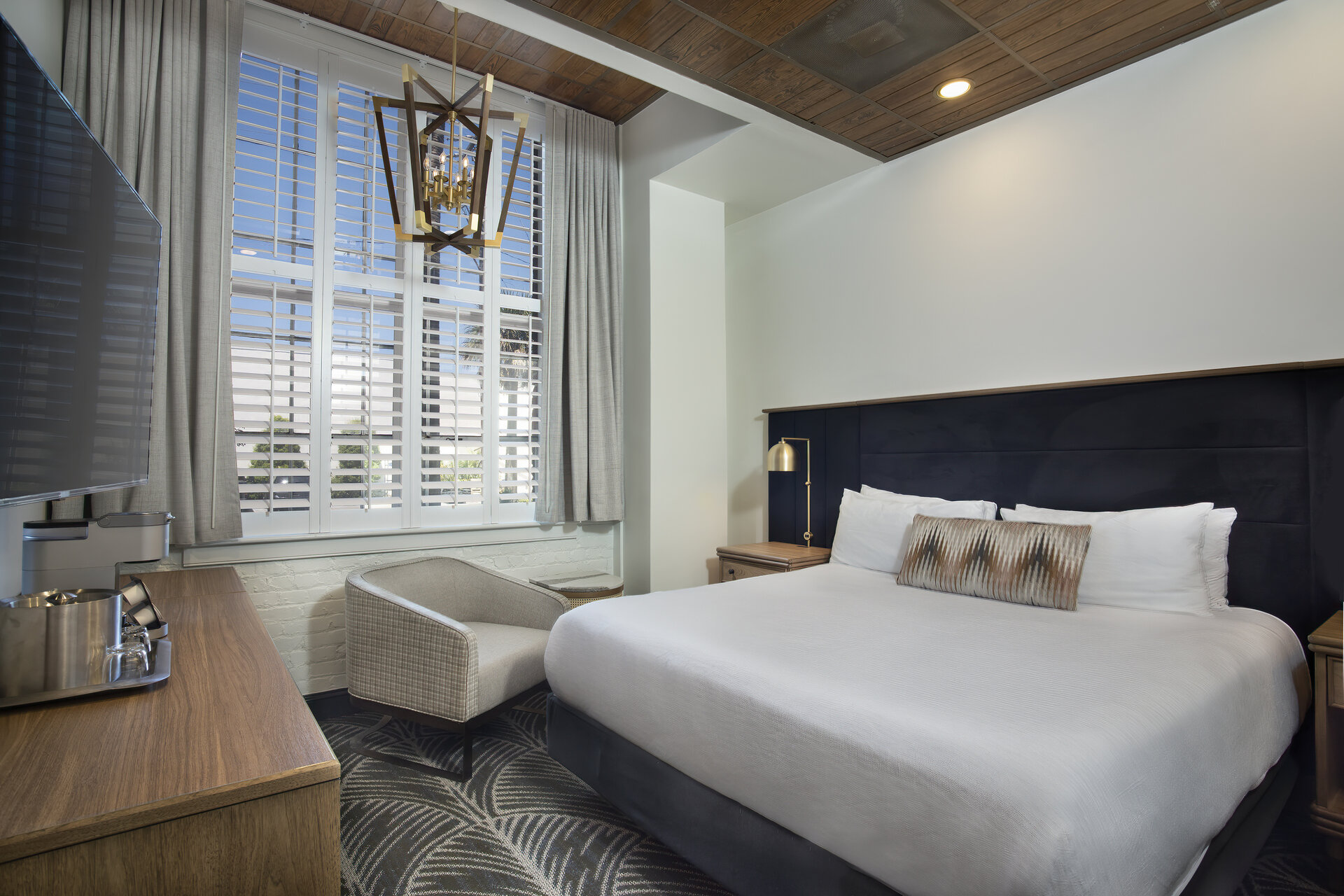 A hotel room with a king-sized bed, dresser, chair, flat-screen TV, and lamp.
