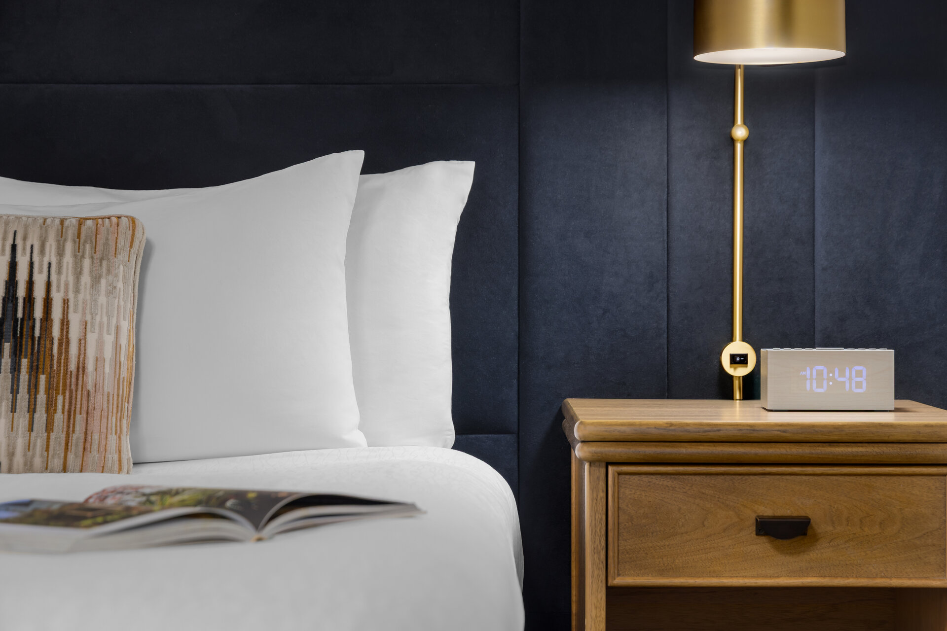 A close-up of a hotel bed with white bedding, a wooden nightstand, a gold lamp, and a digital alarm clock.